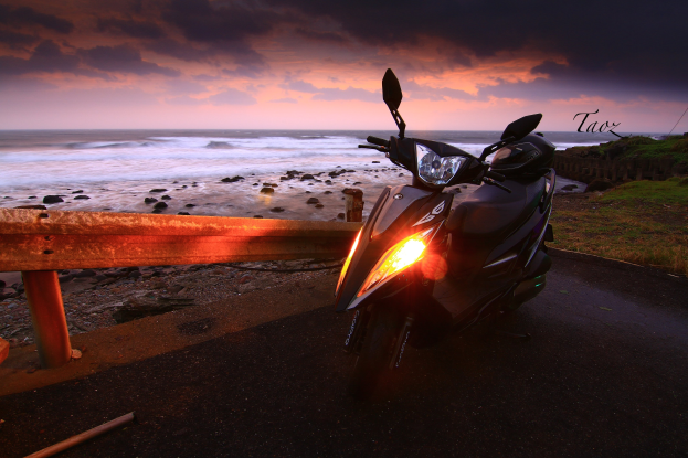 Ein Roller neben einer Straße mit Meerblick, mit einem Zaun, Gras, Steinen und bewölktem Himmel im Hintergrund sowie einem Wasserzeichen.