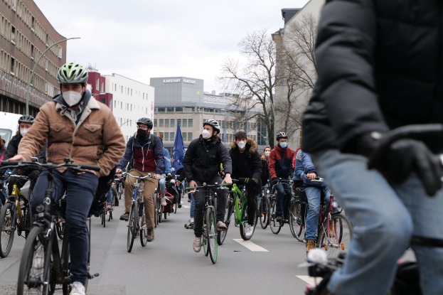 Eine Gruppe von Radfahrern mit Helmen und Handschuhen fährt eine von Bäumen gesäumte Straße in Berlin entlang, mit Gebäuden und geparkten Fahrzeugen im Hintergrund.