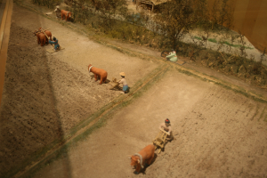 Eine Modellhofszene mit arbeitenden Menschen, Tieren, Pflanzen, Bäumen und einer umgebenden Wand.