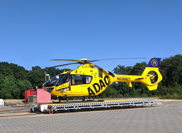 Gelber Hubschrauber auf einem Flughafenvorfeld mit Bäumen und einem klaren blauen Himmel, wahrscheinlich ein Luftrettungshubschrauber in Deutschland mit dazugehöriger Ausrüstung in der Nähe.