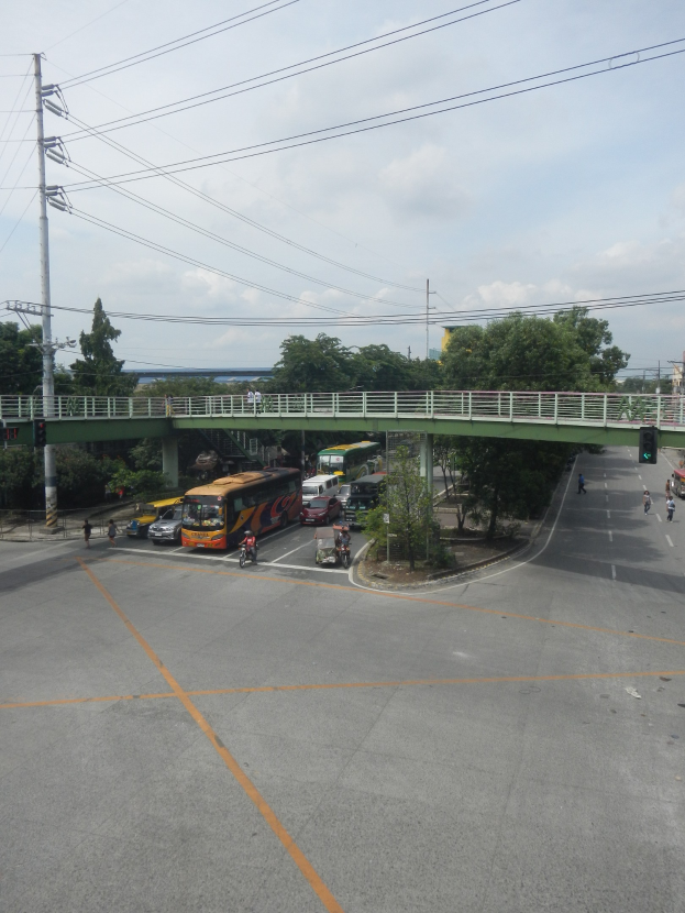Eine Stadtstraße mit Fahrzeugen, eine Brücke mit Geländern und Säulen, Strommasten mit Drähten, Bäume und ein bewölkter Himmel im Hintergrund.