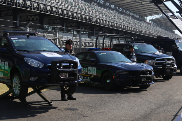 Polizeiautos vor einer Rennstrecke geparkt, mit uniformierten Personen, Stadion und Absperrung im Hintergrund.