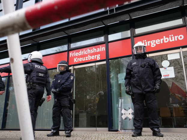 Eine Gruppe von Polizisten in Helmen und Handschuhen steht vor einem Glasgebäude mit einer Stange links und Schildern im Hintergrund, die "Friedrichshain Kreuzberg" und Papiere an den Glaswänden zeigen.