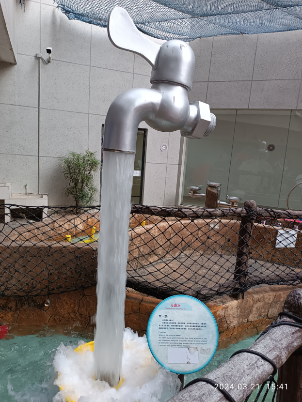 Ein Wasserhahn mit fließendem Wasser vor einem Gebäude mit einem Zaun und einem Schild, mit einer Pflanze und einer Wand im Hintergrund.