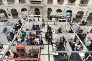 Große Gruppe von Menschen auf einer internationalen Fachmesse mit Ständen, Bannern, Tischen, Stühlen und Luftballons in einem Raum mit architektonischen Merkmalen wie Bögen, Säulen und Geländern.