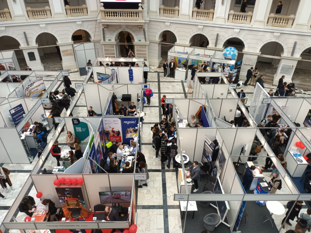 Große Gruppe von Menschen auf einer internationalen Fachmesse mit Ständen, Bannern, Tischen, Stühlen und Luftballons in einem Raum mit architektonischen Merkmalen wie Bögen, Säulen und Geländern.
