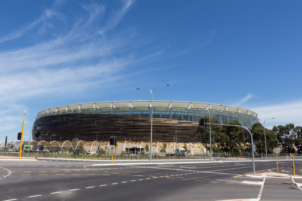 Ein großes Stadion mit starkem Verkehr davor, umgeben von Straßenlaternen, Schildern, Ampeln, Geländern, Bäumen und einem bewölkten Himmel.