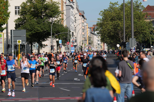 Eine große Gruppe von Läufern nimmt an einem Marathon auf einer Straße teil, die mit Verkehrskegeln, Laternen, Ampeln, Verkehrszeichen, Bäumen, Gebäuden und einem klaren blauen Himmel gesäumt ist.