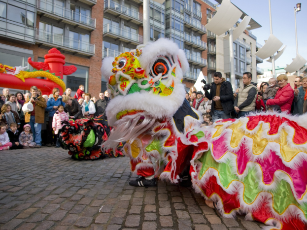 Ein farbenfrohes chinesisches Neujahrsfest in Amsterdam mit einem Löwen tanzen vor einer Zuschauermenge, darunter einige, die das Ereignis fotografieren, vor einer Kulisse aus Gebäuden, Laternenmasten und einem klaren blauen Himmel.