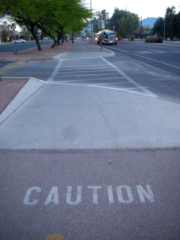 Stadtstraße mit fahrenden Fahrzeugen, ein Gehweg mit einem Warnschild neben einem Baum, Strommasten, Straßenlaternen, Bauzäune, Grünflächen, Gebäude, entfernte Berge und einen klaren blauen Himmel.