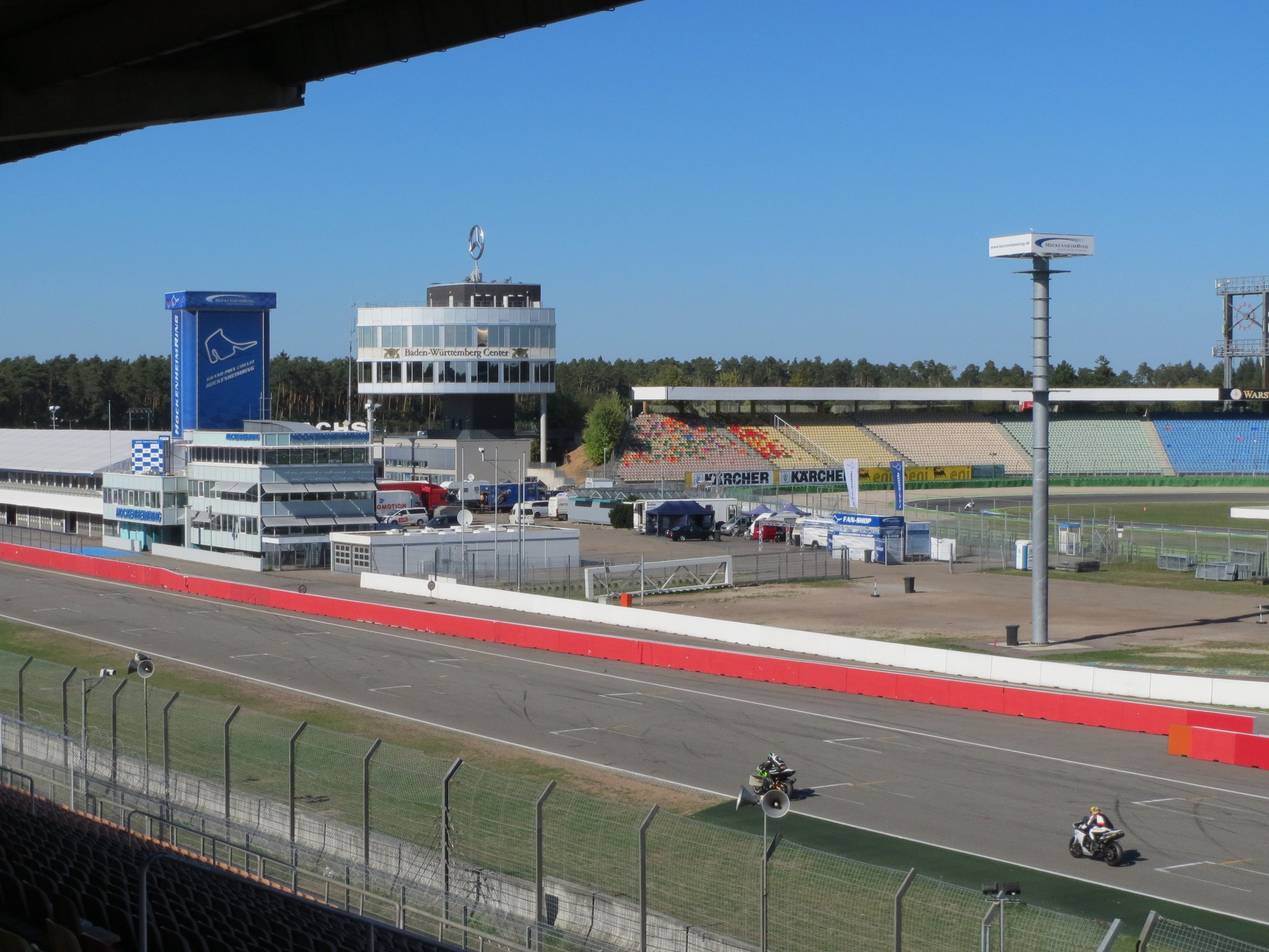Gruppe von Motorradfahrern auf einer Rennstrecke, umgeben von einem Metallzaun, Gras, Fahrzeugen, einem Gebäude, einem Schild, Pfosten, Stadion, Deckenleuchten, Bäumen und einem bewölkten Himmel.