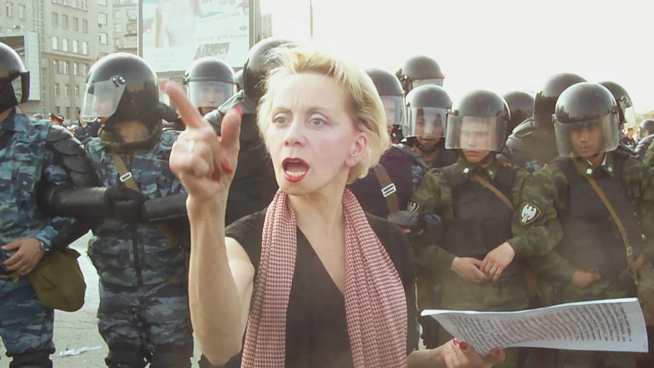 Eine Frau steht einer Reihe von Bereitschaftspolizisten in Helmen gegenüber und hält ein Papier in der Hand, mit Gebäuden, einer Werbetafel und einem blauen Himmel im Hintergrund.
