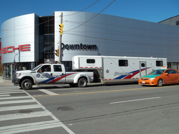 Polizeifahrzeug vor einem Gebäude mit sichtbarem Text geparkt, Verkehrssignal mit Stange und Drähten im Vordergrund, Bäume und klarer blauer Himmel im Hintergrund.