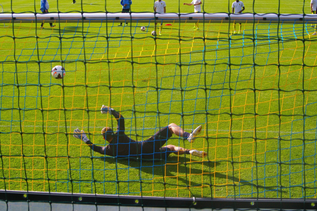 Ein Fussballtorhüter beim Abwehrversuch mit einem Netz im Vordergrund und einigen Personen im Hintergrund, sowie mehreren Fussbällen auf dem Boden.