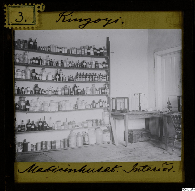 Schwarz-weiß-Foto eines alten Apothekeninneren mit Regalen voller Flaschen, einem Tisch mit Gegenständen, einem Stuhl, einer Tür und einer Wand, mit Text unten.