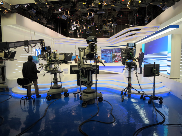 Ein TV-Studio mit mehreren Kameras auf dem Boden, einer sitzenden Person im Vordergrund und einigen stehenden Personen im Hintergrund, mit an der Decke montierten Lampen für eine Livesendung.