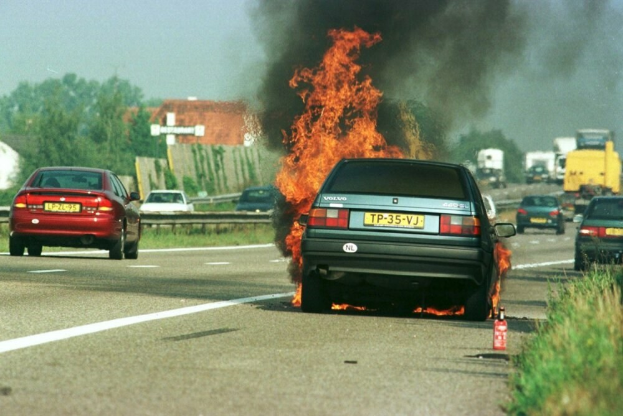 Ein Auto, das in Flammen steht, umgeben von anderen Fahrzeugen, mit Bäumen, Gebäuden und einem klaren blauen Himmel im Hintergrund und einem Feuerlöscher auf der rechten Seite.