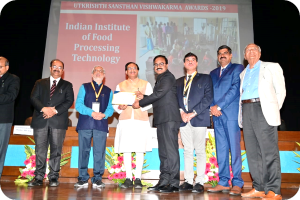 Eine Gruppe von Männern auf einer Bühne mit einem Zertifikat und einem Bildschirm im Hintergrund, der 'Indian Institute of Food Processing Technology Awards 2019' und Blumenschmuck zeigt.