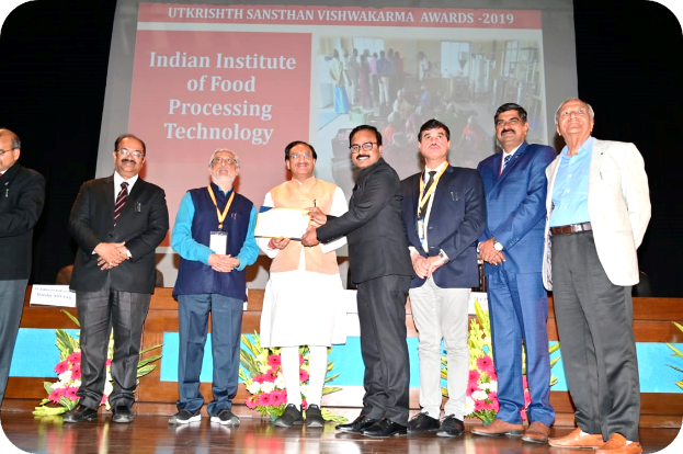 Eine Gruppe von Männern auf einer Bühne mit einem Zertifikat und einem Bildschirm im Hintergrund, der 'Indian Institute of Food Processing Technology Awards 2019' und Blumenschmuck zeigt.