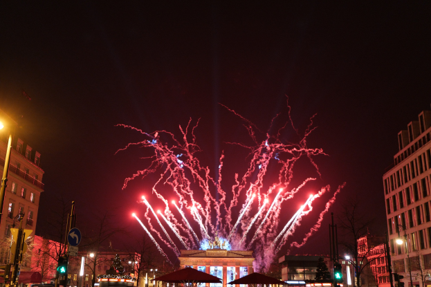 Eine belebte Stadtstraße in Berlin an Silvester, voller Menschen, Fahrzeuge und festlicher Dekoration, mit Feuerwerk, das den Himmel über den Gebäuden erhellt.