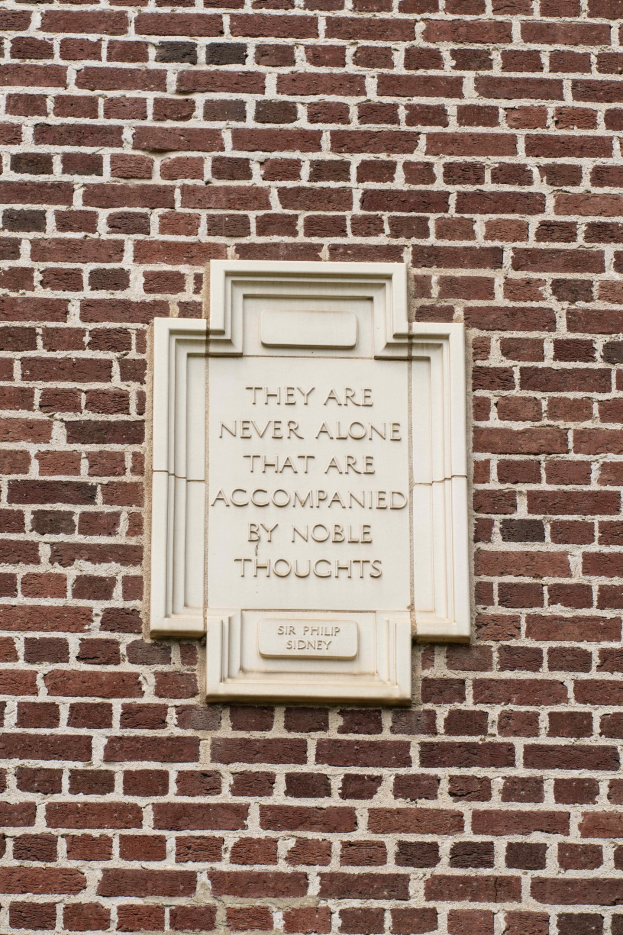 Steintafel an einer Backsteinwand mit der eingravierten Inschrift "Sie sind nie allein, die von edlen Gedanken begleitet sind" in fetter Schrift.