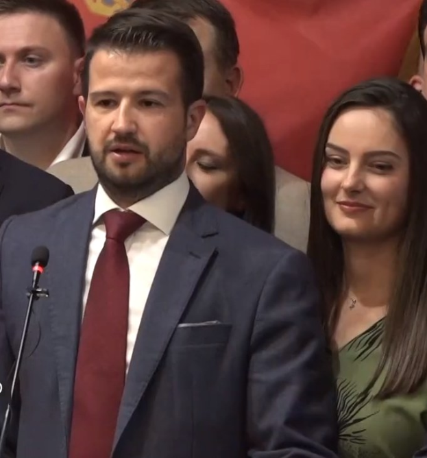 Ein Mann in Anzug und Krawatte spricht in ein Mikrofon an einem Rednerpult, mit einem Banner dahinter und einer Menge davor.