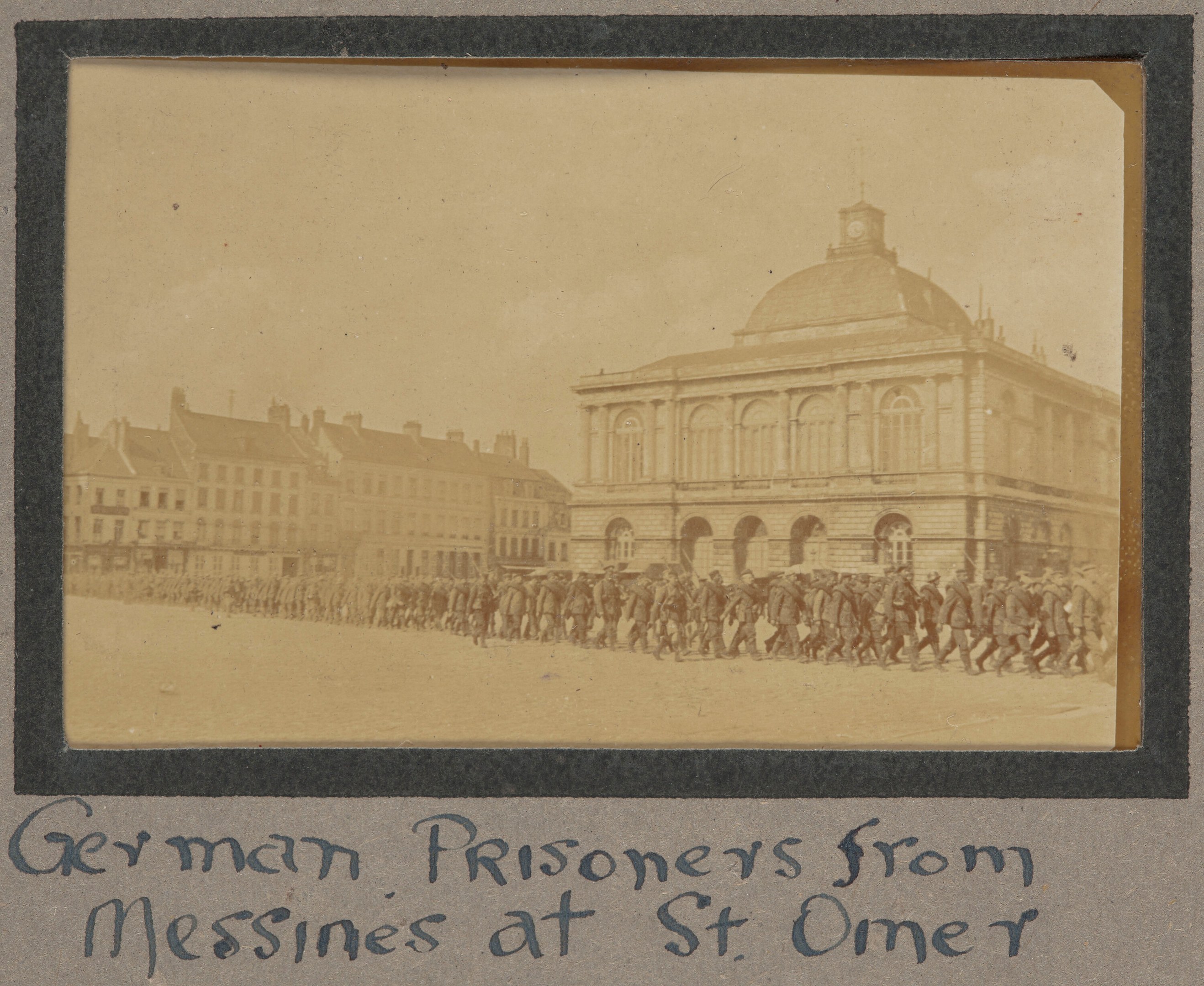 Deutsche Kriegsgefangene aus Messines vor einem Gebäude mit einem klaren blauen Himmel im Hintergrund, unten ist Text sichtbar.