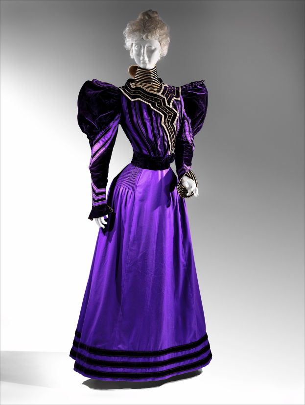 Eine Schaufensterpuppe in einem bodenlangen lila Abendkleid mit einem schwarzen und goldenen gemusterten Rock, der goldene Stickerei und Perlen enthält, einem eng anliegenden Oberteil mit Spitzenbesatz und einem eleganten Haarstil mit zarten Ohrringen.
