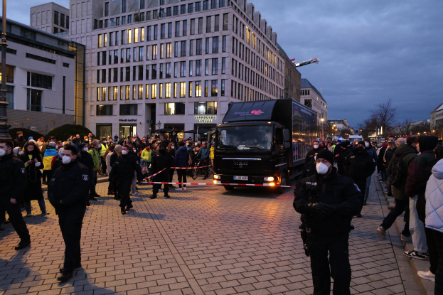 Eine Gruppe von Menschen steht vor einem Lastwagen auf einer Straße, umgeben von Gebäuden, Laternen, Bäumen und einem bewölkten Himmel, wobei einige Mützen und Masken tragen und ein Band mit einem Pfahl im Vordergrund zu sehen ist.