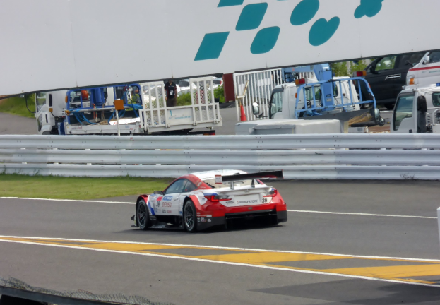 Ein Porsche 911 GT3 RSR auf einer Rennstrecke mit umliegenden Elementen wie Gras, Geländern, Fahrzeugen, Menschen, Bäumen und einer Tafel mit Text oben.