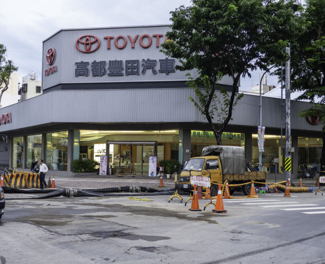 Ein Toyota-Händler in Tokio, Japan, mit Fahrzeugen, Verkehrskegeln, Rohren, Menschen, Bäumen, Pfählen, Brettern und Gebäuden im Vordergrund und einem bewölkten Himmel im Hintergrund.