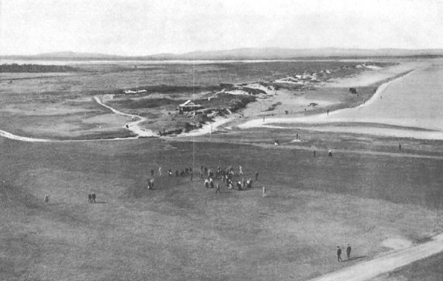 Schwarz-weiß-Foto von Golfern auf dem 18. Loch des Royal Birkham Golf Clubs mit saftig grünem Rasen, verstreuten Häusern, sanften Hügeln und einem strahlend blauen Himmel.
