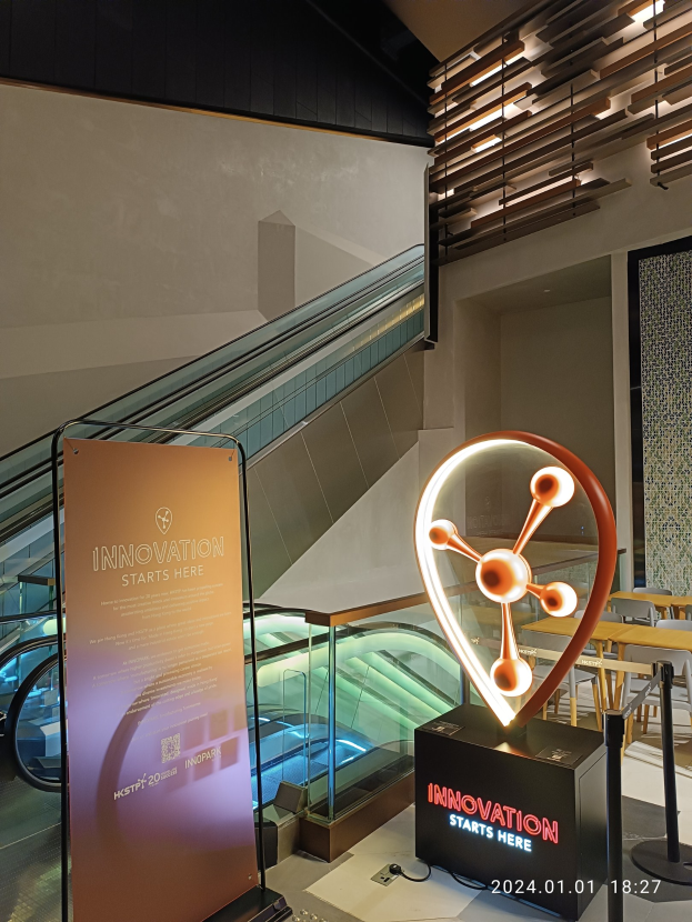 Großes Schild in einem Gebäude mit der Aufschrift "Innovation Starts Here" und einer Rolltreppe im Hintergrund, Tische und Stühle auf der rechten Seite und eine Tafel auf der linken Seite, die die Teilnahme an der HSTP20 Expo anzeigt.