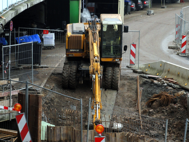 Eine Baustelle mit einem großen Bagger in der Mitte, umgeben von einem Zaun, Schildern, Pfählen und verschiedenen Gegenständen, mit Fahrzeugen auf einer Straße und einer Brücke im Hintergrund.