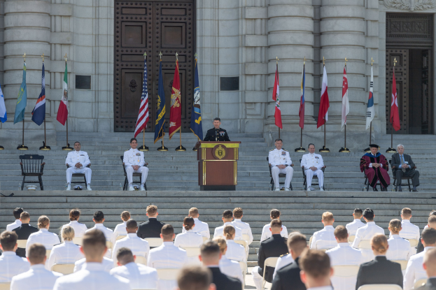 Gruppe von Menschen in weißen Uniformen auf Treppe eines Gebäudes mit Podium und Flaggen vor einem großen Gebäude mit Türen und Säulen.
