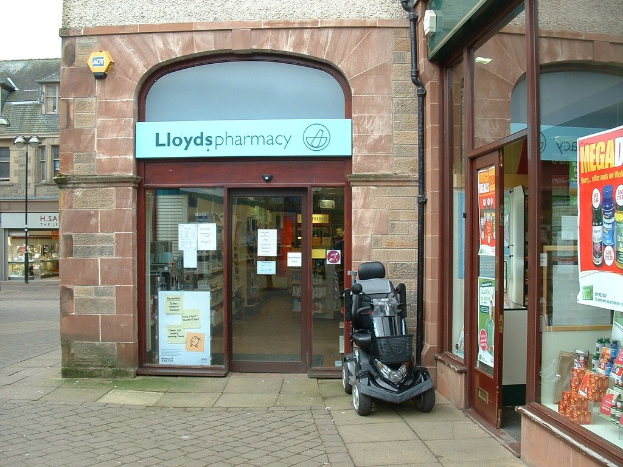 Apothekenladen mit einem vor ihm geparkten Fahrzeug und einem Gebäude in der linken Ecke.