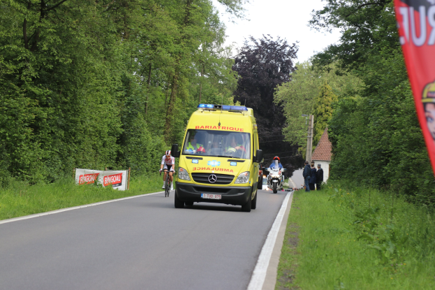 Ambulanz fährt auf Straße mit Radfahrern daneben, gesäumt von Gras, Bäumen, Häusern, Strommasten und einem klaren blauen Himmel.