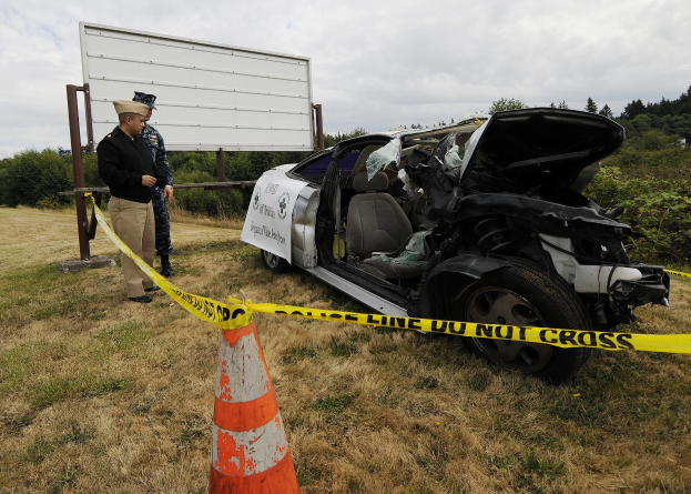Ein Polizeibeamter in Uniform steht neben einem schwer beschädigten Auto am Straßenrand, mit eingedellter Haube und zersplitterten Fenstern, während eine Verkehrskegel und eine Absperrung in der Nähe sichtbar sind.