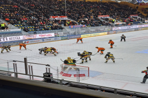 Gruppe von Menschen, die Hockey auf einer Indoor-Eisbahn mit einem Netz spielen, umgeben von einem Zaun, unter Stadionbeleuchtung, mit Zuschauern im Hintergrund und Schildern.
