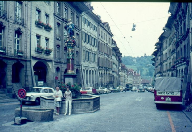 Altes Schwarz-Weiß-Foto einer Stadtstraße mit Fußgängern, parkenden Autos, Gebäuden mit Fenstern, Topfpflanzen, einer Statue auf einem Sockel, einem Schild, einem Fahrzeug auf der Straße, Bäumen und einem klaren blauen Himmel.