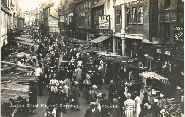 Ein Schwarz-Weiß-Foto eines belebten Straßenmarkts in Croydon, mit Menschen, die an Ständen auf beiden Seiten vorbeigehen, Gebäuden mit Fenstern im Hintergrund und einem Lichtmast.