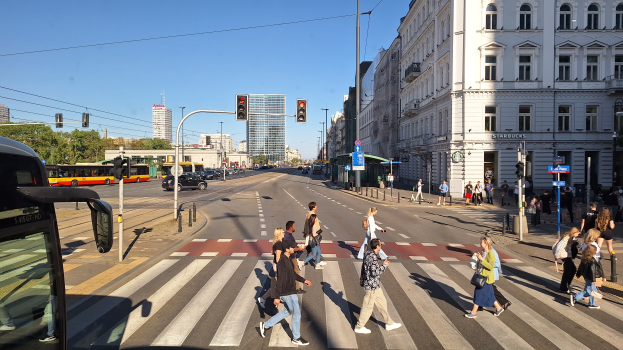 Gruppe von Menschen, die eine Straße an einer Ampel überqueren, mit einem Bus auf der linken Seite, Fahrzeugen auf der Straße, Verkehrszeichen, Laternen, Gebäuden, Bäumen und einem klaren blauen Himmel im Hintergrund.