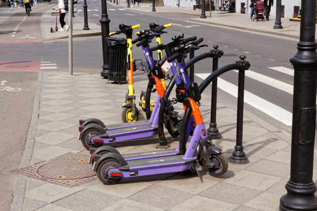 Eine Reihe von Elektroscootern, die entlang einer Straße geparkt sind, mit Fußgängern auf dem Gehweg, Radfahrern, einem Müllcontainer im Vordergrund und Gebäuden, Bäumen und Pflanzen im Hintergrund.