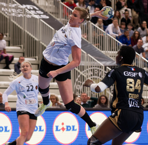 Eine Gruppe von Frauen, die auf einem Platz Handball spielen, mit einem Ball in der Luft, Zuschauern im Hintergrund und einem "Handball-Weltmeisterschaft 2019 - Tag 2"-Schild.