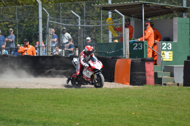 Eine Person auf einem Motorrad fährt über ein grasbewachsenes Feld, mit Zuschauern in Mützen und Schutzbrillen, einem Maschendrahtzaun, Pfählen, Bäumen, Treppen mit Geländern und einem Schuppen mit Nummern und Texttafeln im Hintergrund.