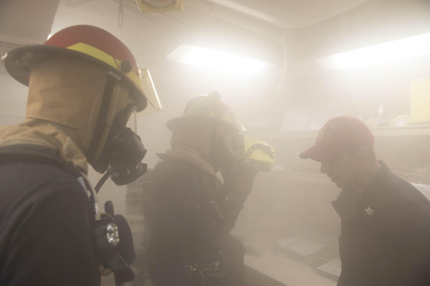 Feuerwehrleute mit Helmen und Rauch aus dem Mund während einer Übung, einer hält eine Kamera, mit Hintergrundlichtern und -ausrüstung.