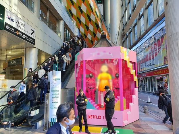 Eine Gruppe von Menschen steht vor einem glaswandigen Gebäude mit Texttafeln, einige tragen Masken, während eine Rolltreppe mit Menschen daneben aufsteigt; ein pinkfarbener Kasten in der Mitte zeigt Schaufensterpuppen und trägt 'Netflix' neue Netflix-Bühne in Tokio'.