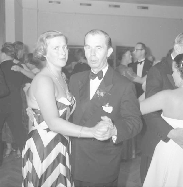 Ein Mann in einem schwarzen Blazer, weißem Hemd und schwarzen Hosen tanzt mit einer Frau in einem unbekannten Outfit auf einer formellen Veranstaltung, indem sie sich an den Händen halten und zur Musik schwenken, während sie in der Nähe einer Wand tanzen.