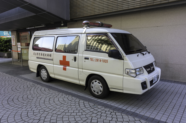 Ambulanz vor einem Gebäude mit einem Schild geparkt, das darauf hinweist, dass es ein Notfahrzeug ist, umgeben von Bäumen und Pflanzen.