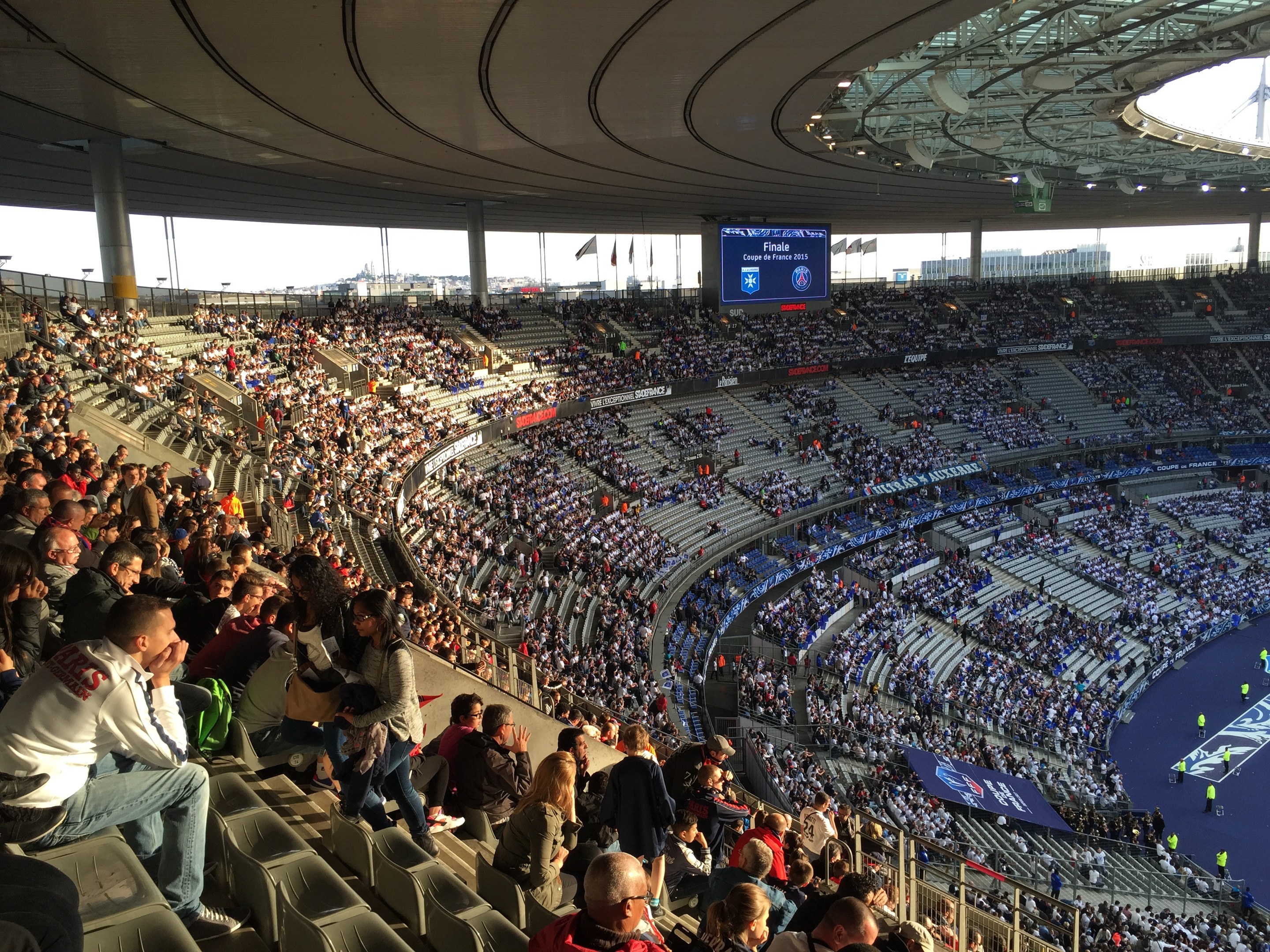 Große Menschenmenge in einem Stadion bei einem Fussballspiel sitzend, mit einer Bühne auf der rechten Seite, Fahnen, Masten, einem Bildschirm und der Allianz Arena in München, Deutschland im Hintergrund.